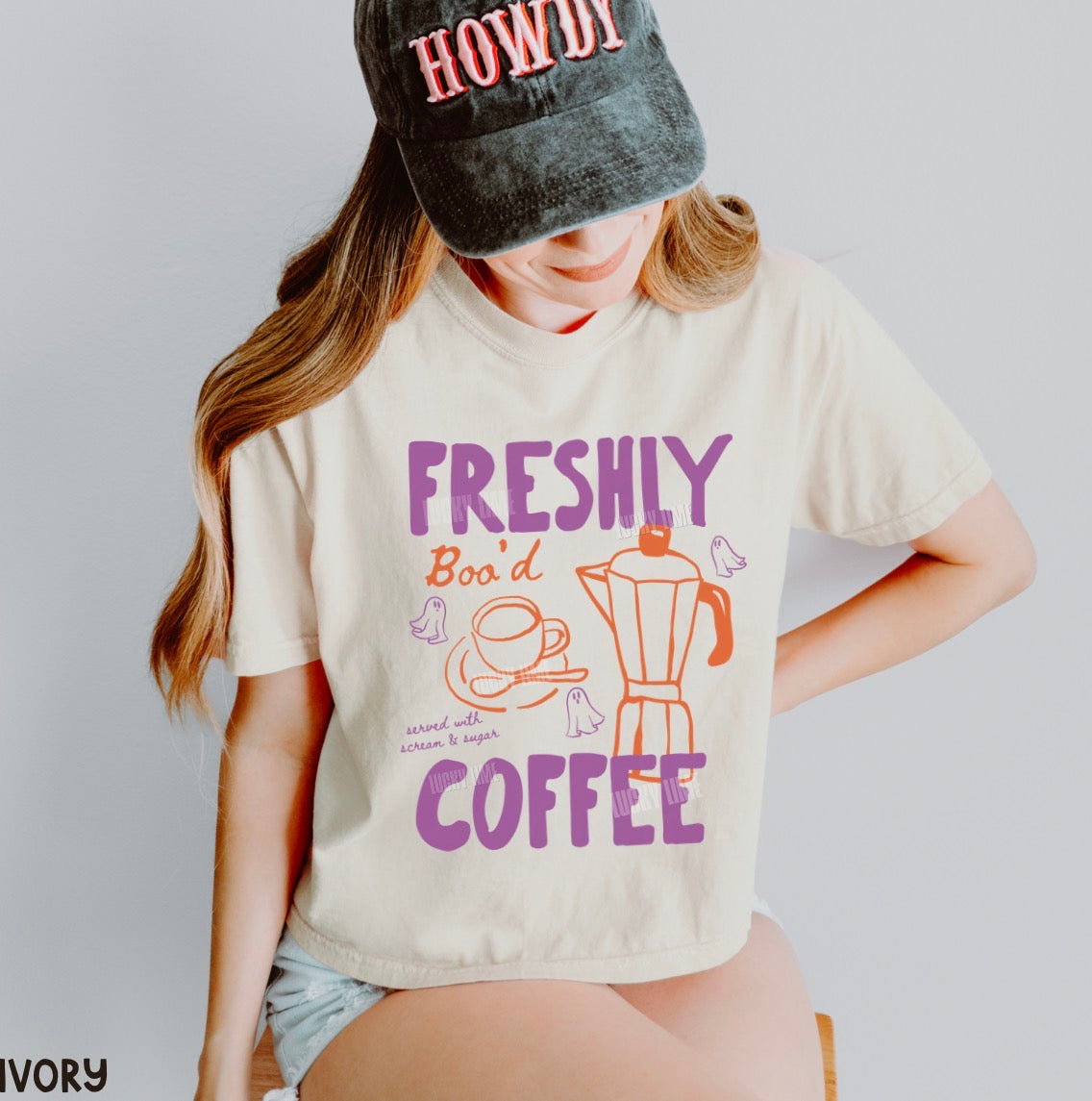 A woman wearing a cream-colored graphic t-shirt with purple and orange text reading 'FRESHLY BOODED COFFEE' and a black cap with 'HOWDY' in red, posed against a light gray background.