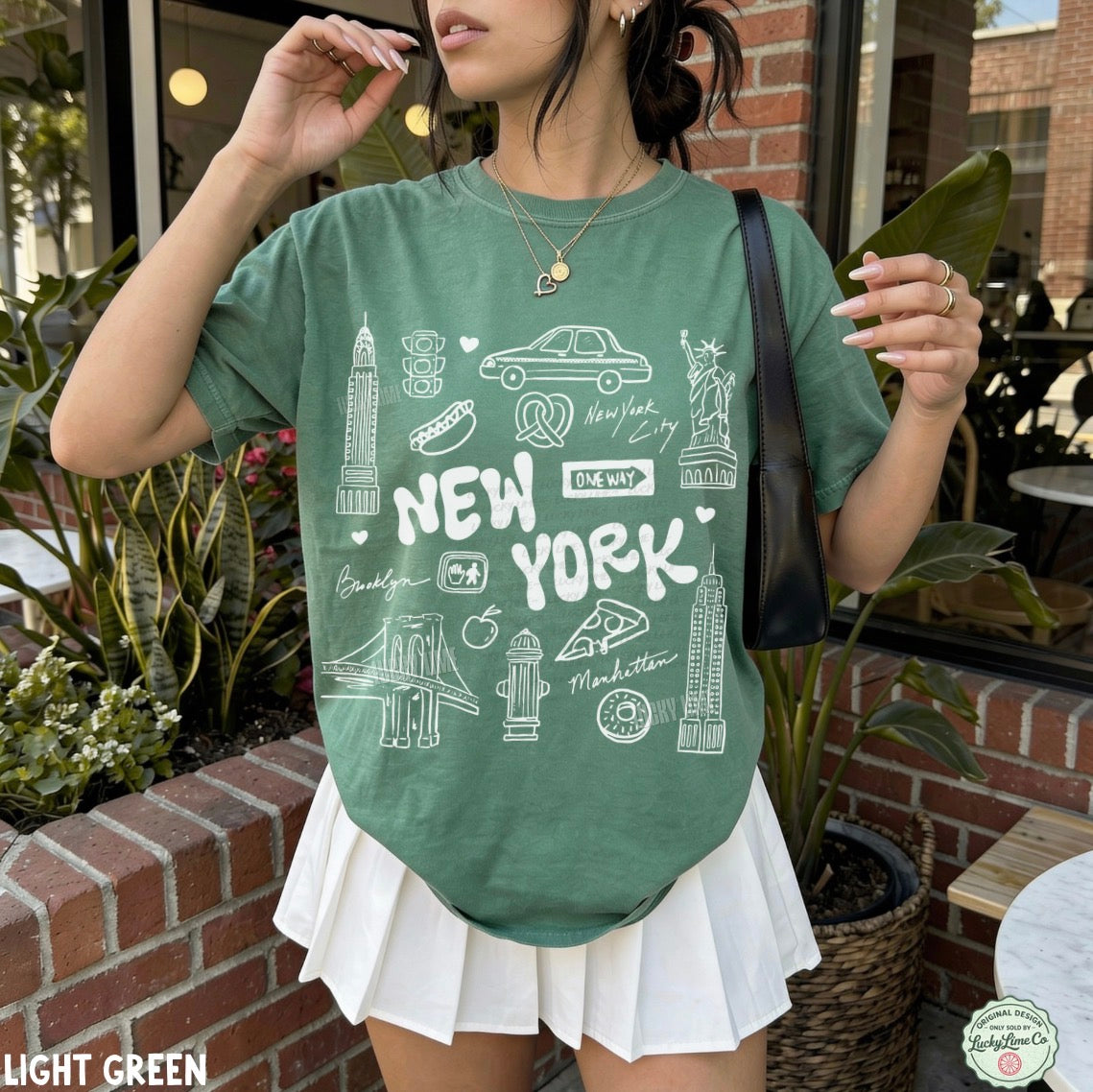A woman wearing a light green graphic t-shirt with white New York City-themed illustrations and text, paired with a white pleated skirt, standing in front of a brick wall and cafe window.