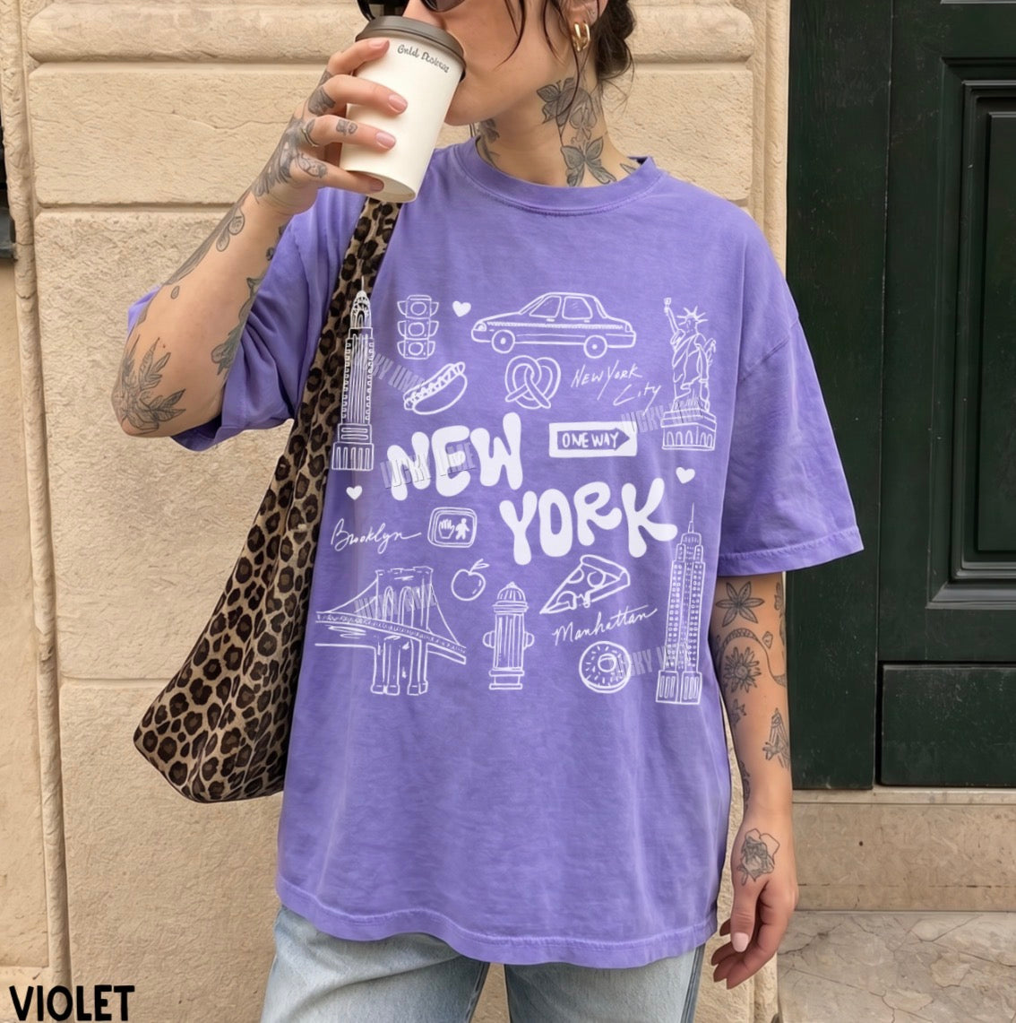A person wearing an oversized purple T-shirt with white New York City-themed graphics and text, standing outdoors against a beige stone wall and holding a coffee cup.