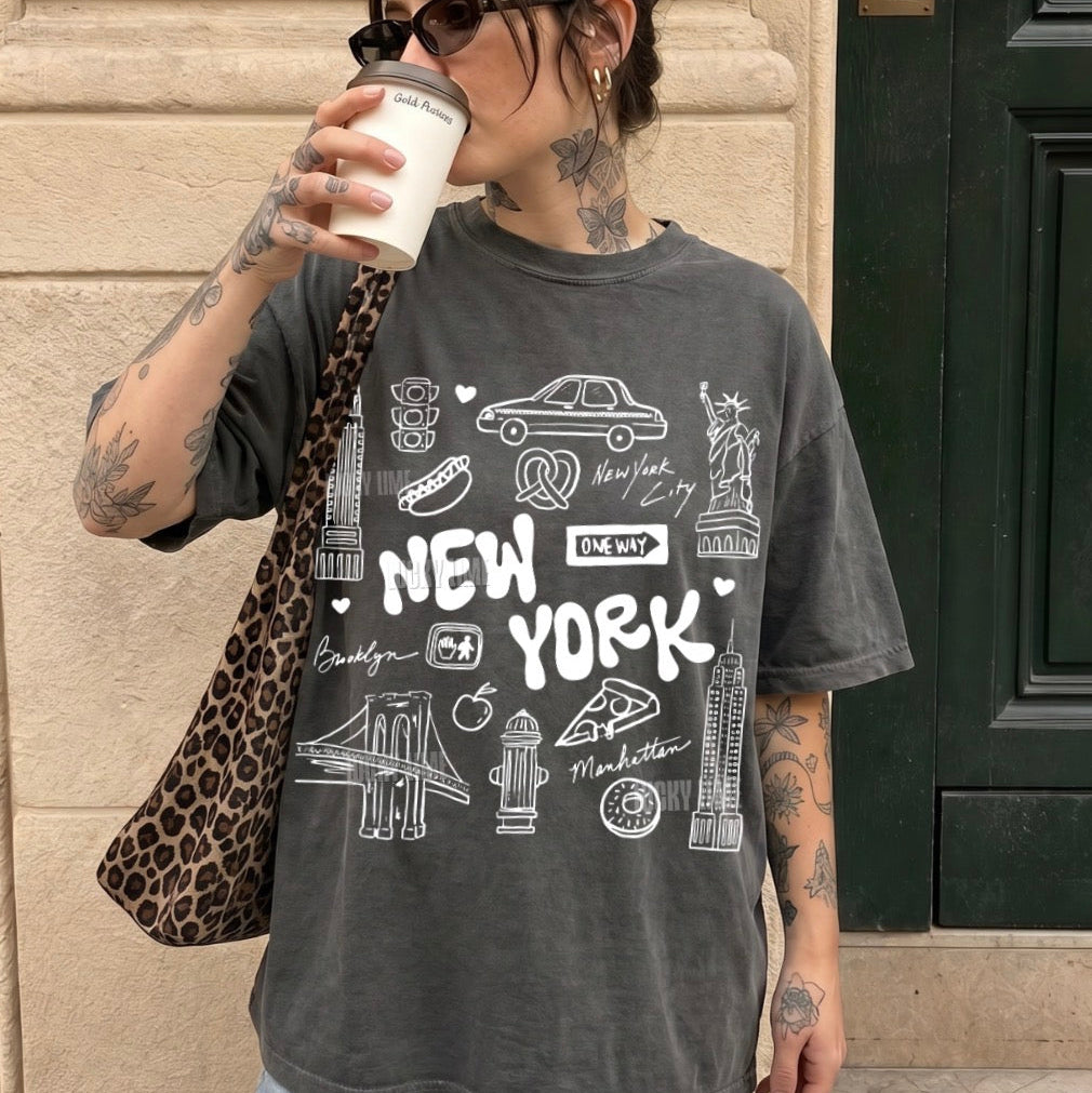 A woman wearing a dark gray acid-wash graphic t-shirt with white New York-themed illustrations and text, holding a coffee cup and wearing a leopard-print shoulder bag, standing outdoors against a beige stone wall.