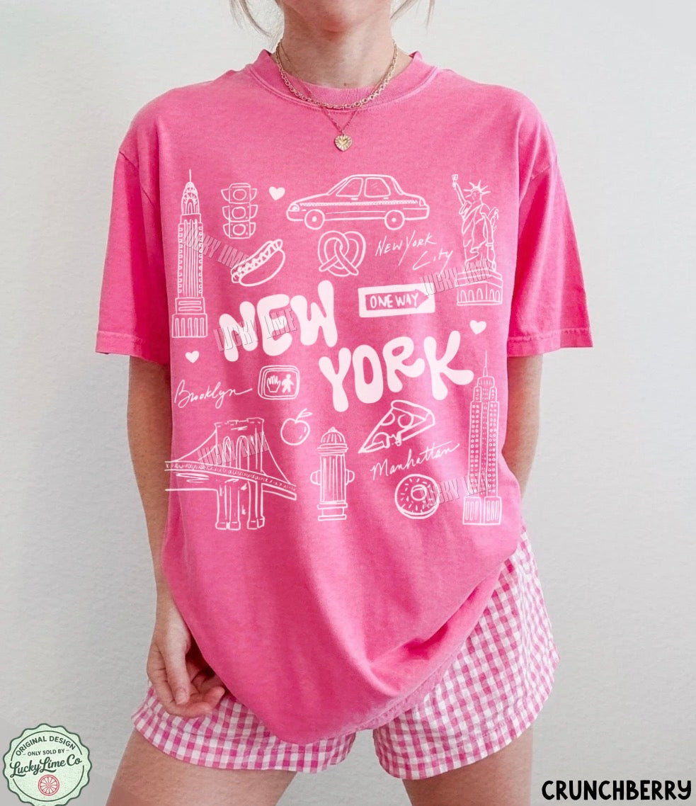 A woman wearing a pink short-sleeve graphic T-shirt with white New York-themed illustrations and text, paired with pink-and-white gingham shorts. The photo is cropped from the neck down against a plain light background, with a small 'CRUNCHBERRY' label in the bottom right corner.