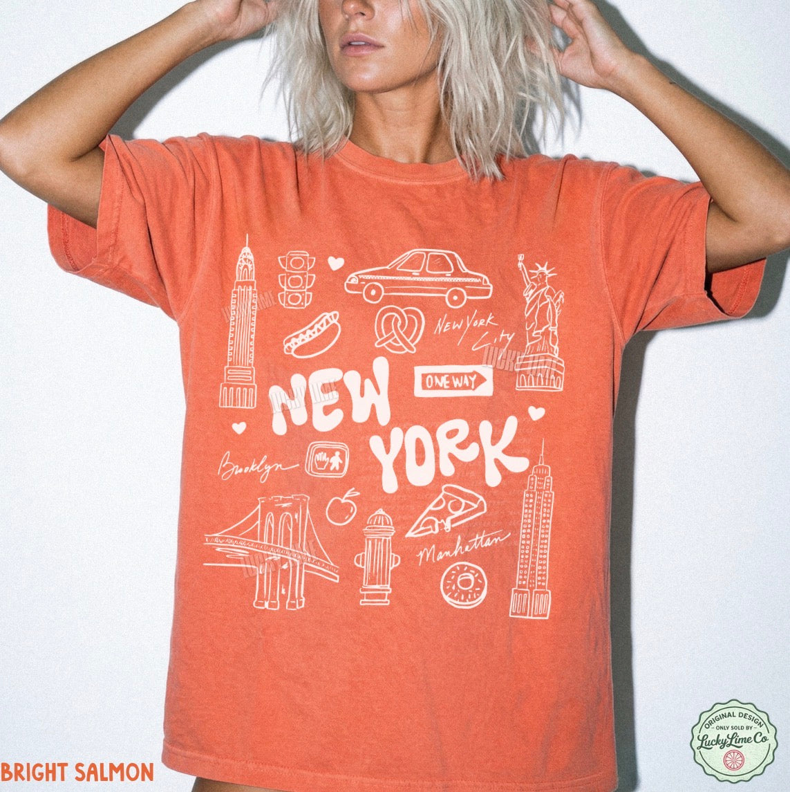 A woman wearing a bright salmon orange graphic t-shirt with white line drawings of New York City landmarks and text, set against a plain white background.