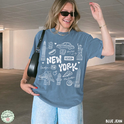 A woman wearing a blue graphic t-shirt with white New York City illustrations, paired with light blue jeans and a black shoulder bag, standing in a parking garage.