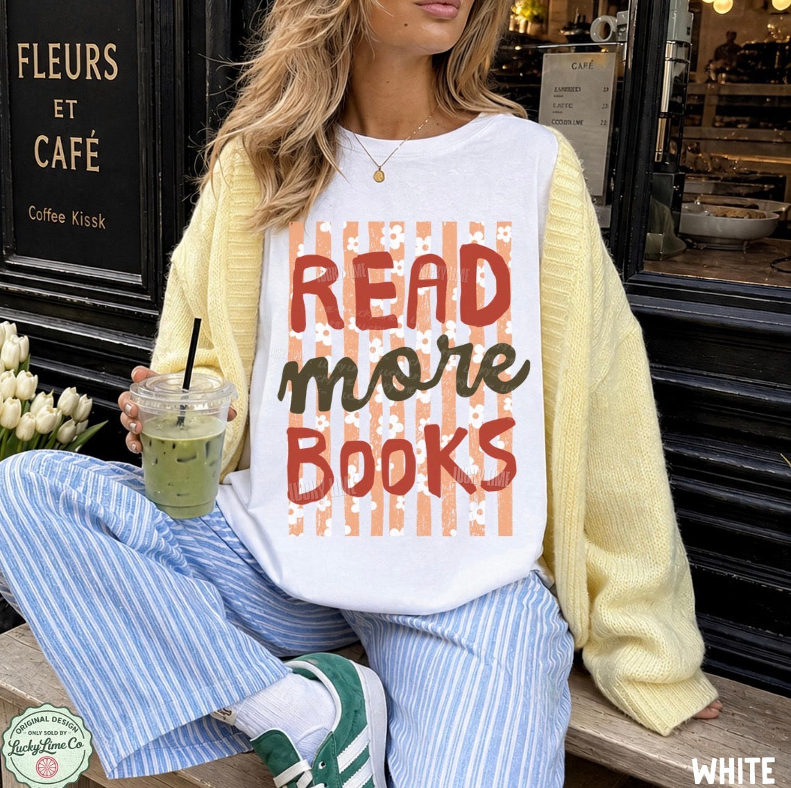 A woman sitting on a bench in front of a coffee shop, wearing a white graphic sweatshirt with yellow and orange striped lettering that reads 'READ more BOOKS'. She has on a yellow cardigan, blue and white striped pants, and green sneakers, and is holding an iced coffee.
