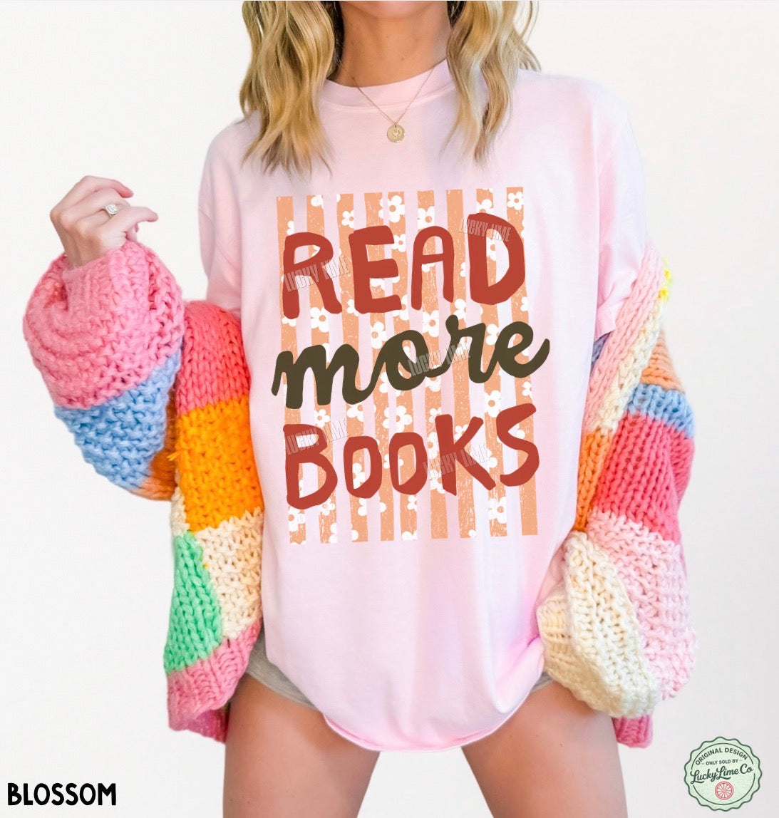 A woman wearing a light pink graphic T-shirt with the words 'READ more BOOKS' in brown and black lettering over a striped and floral pattern. She is holding a chunky, multicolored knit cardigan with pink, blue, orange, yellow, green, and cream sections.