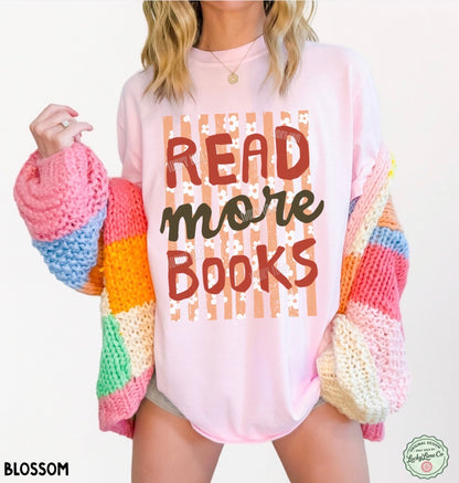 A woman wearing a light pink graphic T-shirt with the words 'READ more BOOKS' in brown and black lettering over a striped and floral pattern. She is holding a chunky, multicolored knit cardigan with pink, blue, orange, yellow, green, and cream sections.