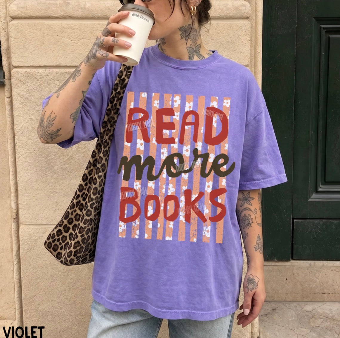 A person wearing an oversized violet graphic T-shirt with the text 'READ more BOOKS' over a striped design, holding a coffee cup and carrying a leopard-print shoulder bag in front of a stone wall and dark door.