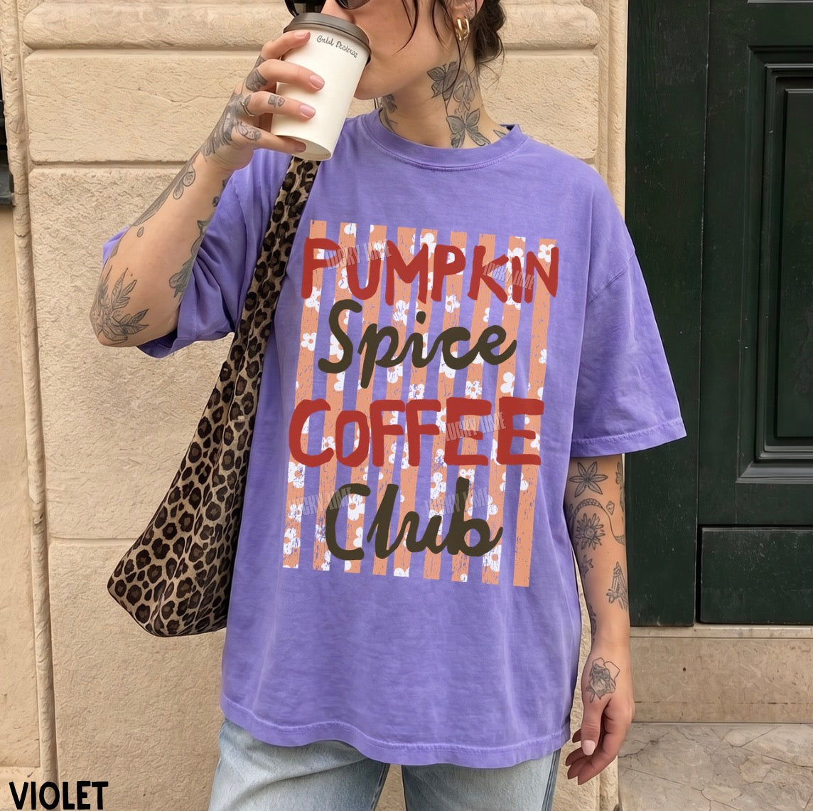 A person wearing an oversized purple graphic T-shirt with the text 'Pumpkin Spice Coffee Club' printed on the front, standing outdoors against a beige stone wall and holding a coffee cup. A leopard-print shoulder bag is also visible.