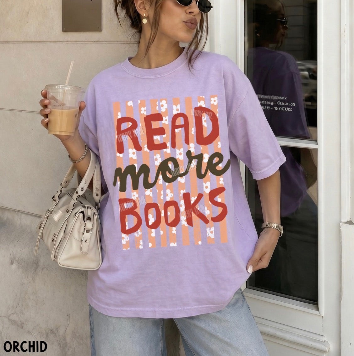 A woman wearing a lavender graphic t-shirt with the text 'READ more BOOKS' in red and dark brown lettering over a peach and white striped background. She is holding a drink and a white handbag, standing in front of a building entrance.