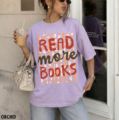 A woman wearing a lavender graphic t-shirt with the text 'READ more BOOKS' in red and dark brown lettering over a peach and white striped background. She is holding a drink and a white handbag, standing in front of a building entrance.