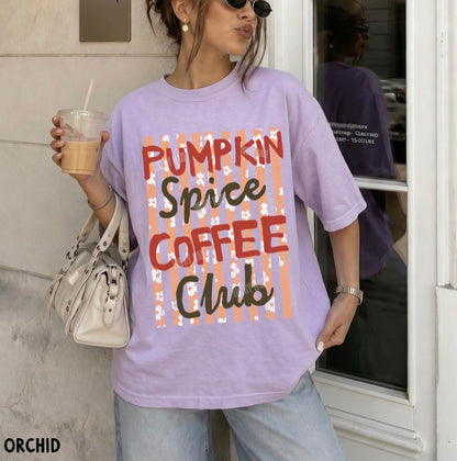 A woman wearing an oversized lavender graphic T-shirt with the text 'Pumpkin Spice Coffee Club' in red and black lettering over a striped and polka-dot background. She is holding an iced coffee and carrying a white handbag, standing in front of a storefront window.