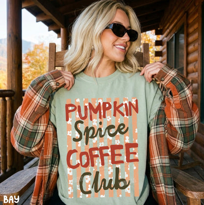 A woman wearing a sage green crewneck sweatshirt with 'PUMPKIN SPICE COFFEE CLUB' printed on the front, styled with an orange and green plaid flannel shirt on a rustic wooden porch.