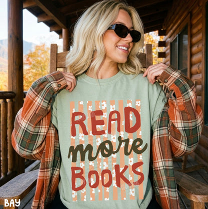 A woman wearing a sage green graphic sweatshirt with the text 'READ more BOOKS' in red and brown lettering, styled with an orange and green plaid flannel shirt on a rustic wooden porch.