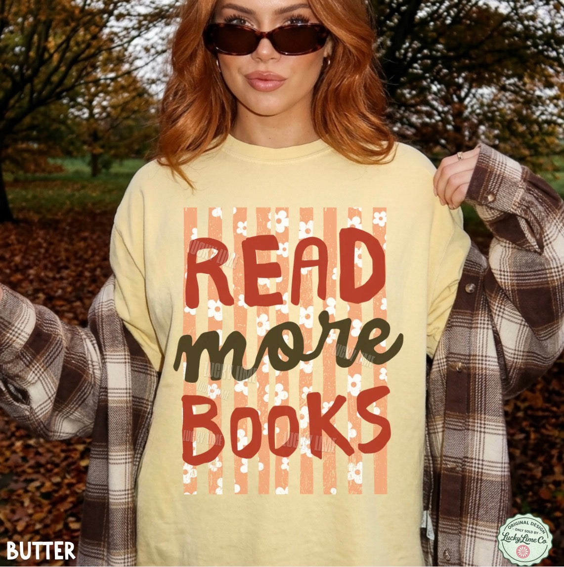 A woman outdoors wearing a pale yellow graphic T-shirt that reads 'READ more BOOKS' over vertical peach stripes, with a brown plaid shirt draped off her shoulders. Trees and fallen leaves are visible in the background.