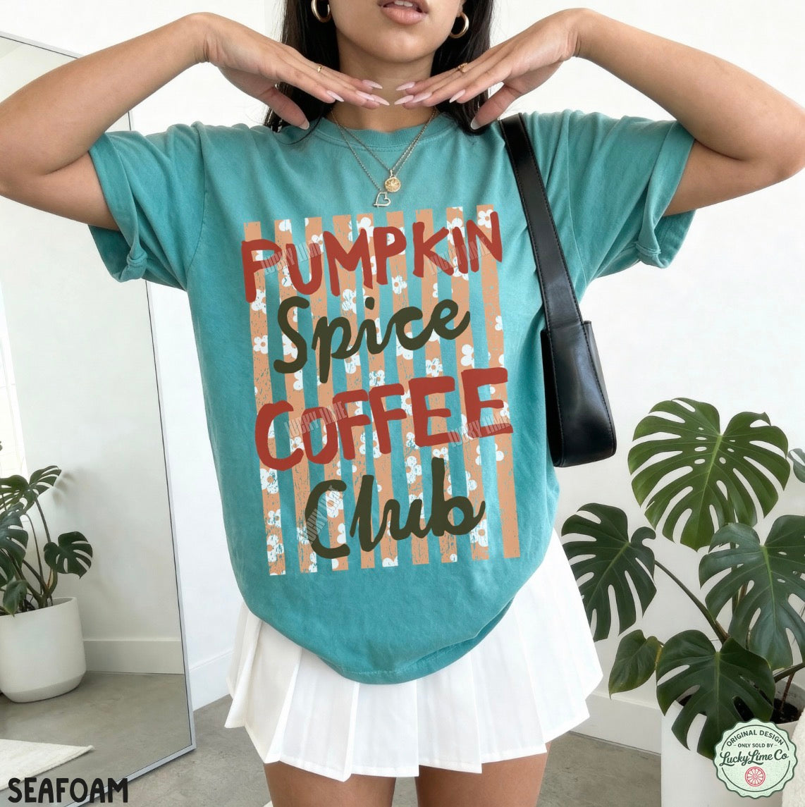 A woman wearing a teal graphic t-shirt with the text 'PUMPKIN SPICE COFFEE CLUB' over a striped design, paired with a white pleated skirt and a black shoulder bag, in a brightly lit indoor setting with potted plants.
