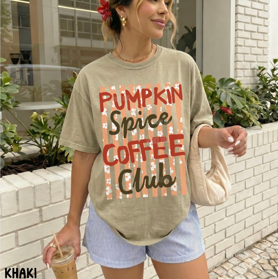 A woman wearing an oversized beige graphic T-shirt that reads 'PUMPKIN SPICE COFFEE CLUB' in red and dark brown lettering over a striped pattern, paired with light blue striped shorts and holding an iced coffee in an outdoor patio setting.