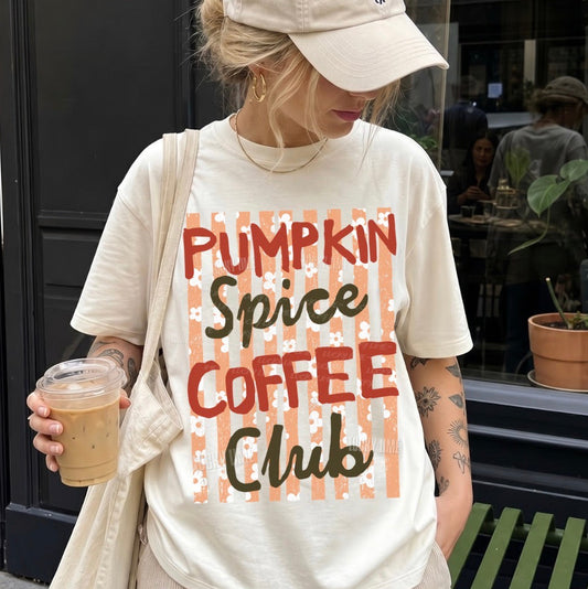 A woman wearing a cream-colored graphic t-shirt with 'PUMPKIN Spice COFFEE Club' printed on the front, paired with a matching beige baseball cap and holding an iced coffee in front of a storefront.