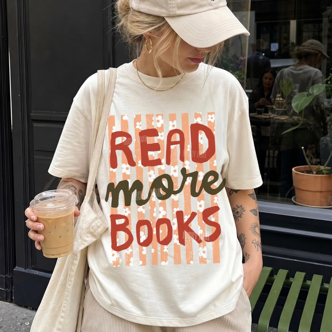 A woman wearing a cream-colored graphic t-shirt with the text 'READ MORE BOOKS' in red and dark green lettering over a peach striped background, paired with a matching beige cap and holding an iced coffee on a city street.