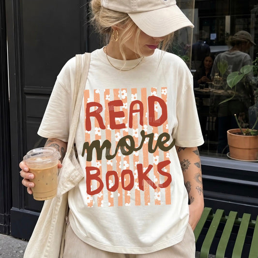 A woman wearing a cream-colored graphic t-shirt with the text 'READ MORE BOOKS' in red and dark green lettering over a peach striped background, paired with a matching beige cap and holding an iced coffee on a city street.