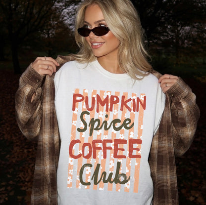 A woman outdoors wearing a white graphic sweatshirt that reads 'Pumpkin Spice Coffee Club' in red and dark green lettering over peach vertical stripes, with a brown plaid flannel shirt draped off her shoulders.