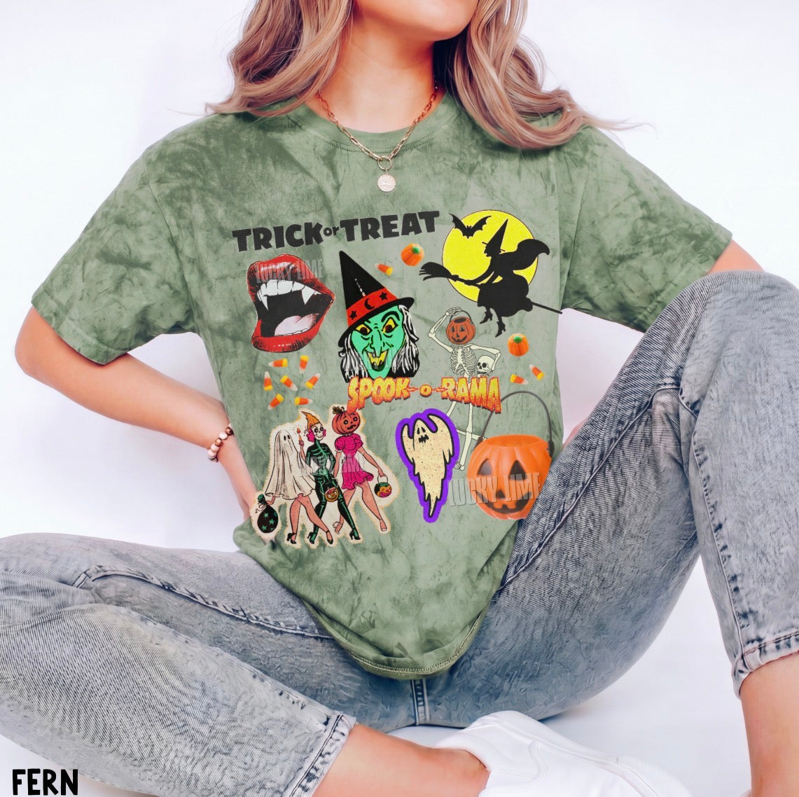 A woman wearing a faded olive green short-sleeve t-shirt with a colorful Halloween graphic print that reads 'TRICK or TREAT' and features spooky illustrations, paired with gray jeans.