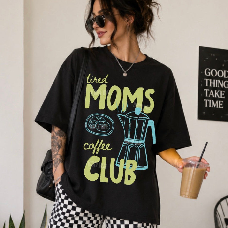 A woman wearing a black graphic t-shirt with the text 'tired MOMS coffee CLUB' in yellow-green and blue, paired with black and white checkered pants, holding an iced coffee in a room setting.