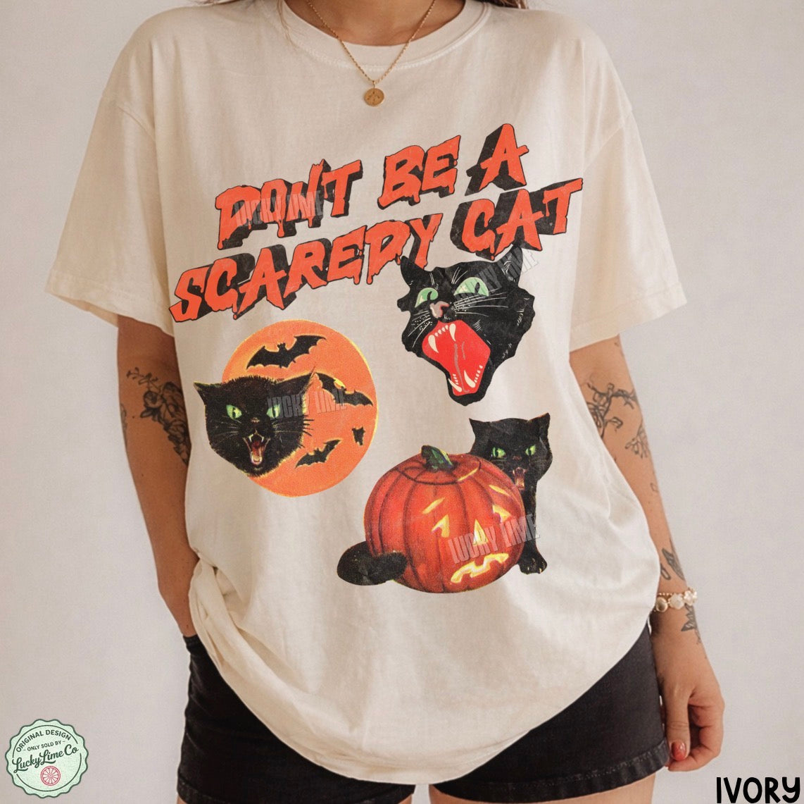 A person wearing an oversized ivory graphic T-shirt with a Halloween design featuring black cats, bats, a jack-o'-lantern, and the text 'DON'T BE A SCAREY CAT' in orange and black. The photo is cropped from the neck down against a plain light background.