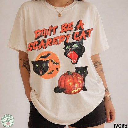 A person wearing an oversized ivory graphic T-shirt with a Halloween design featuring black cats, bats, a jack-o'-lantern, and the text 'DON'T BE A SCAREY CAT' in orange and black. The photo is cropped from the neck down against a plain light background.