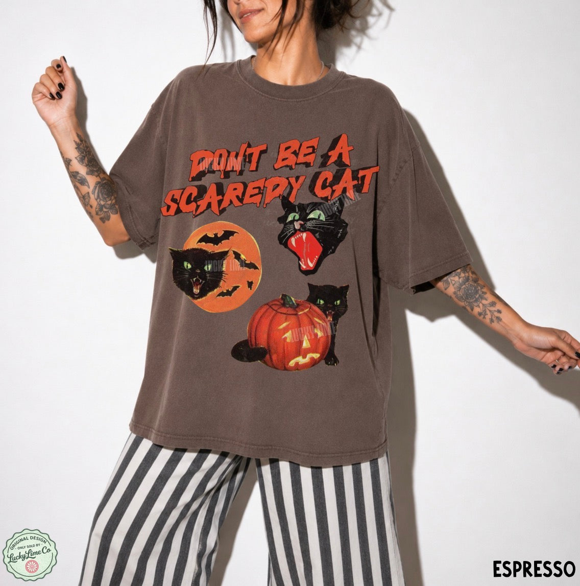 A person wearing an oversized brown graphic T-shirt with the phrase “DON’T BE A SCAREDY CAT” and Halloween-themed cat illustrations, paired with black-and-white striped pants, against a plain light background.