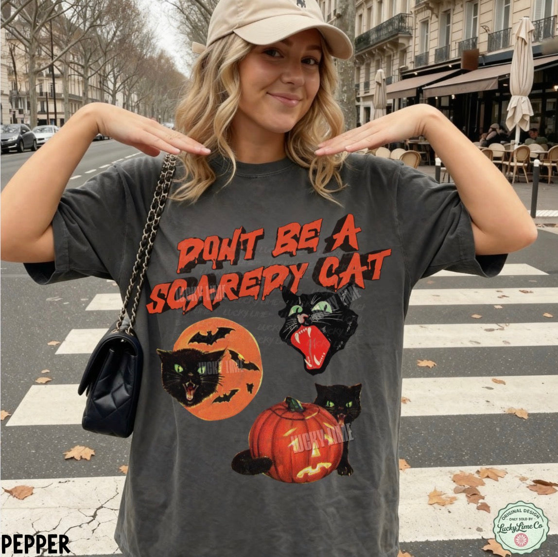 A woman wearing a dark gray graphic t-shirt with a Halloween design that reads 'DON'T BE A SCAREDY CAT' above three black cats, one on an orange moon, one with its mouth open, and one on a pumpkin. She is posing on a city street crosswalk.