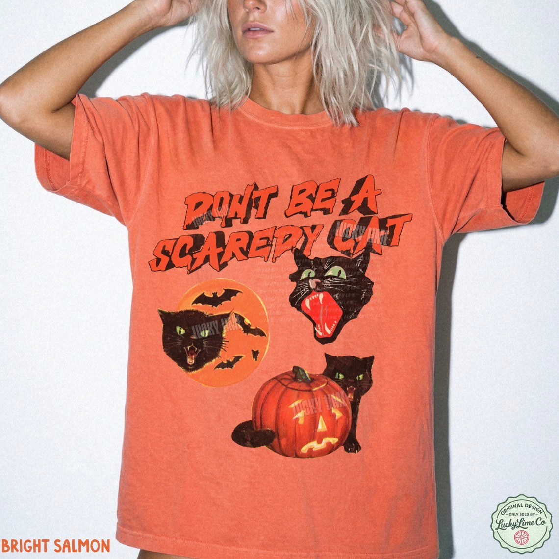 A woman wearing a bright salmon orange graphic t-shirt with a Halloween theme featuring black cats, a jack-o'-lantern, and the text 'DON'T BE A SCAREY CAT'.