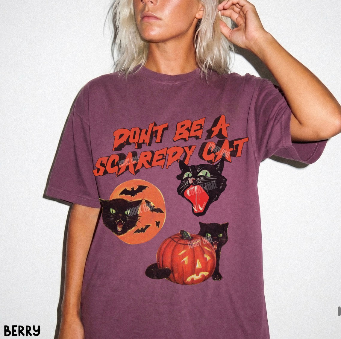 A model wearing a berry purple short-sleeve t-shirt with a Halloween graphic that reads 'DON'T BE A SCAREDY CAT' above three black cats, a jack-o'-lantern, and bats on an orange background.