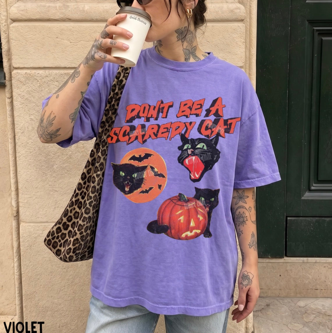 A person wearing an oversized purple graphic T-shirt with the text 'DON'T BE A SCAREY CAT' and Halloween-themed cat illustrations, paired with light blue jeans and a leopard-print shoulder bag, standing outdoors against a beige wall.