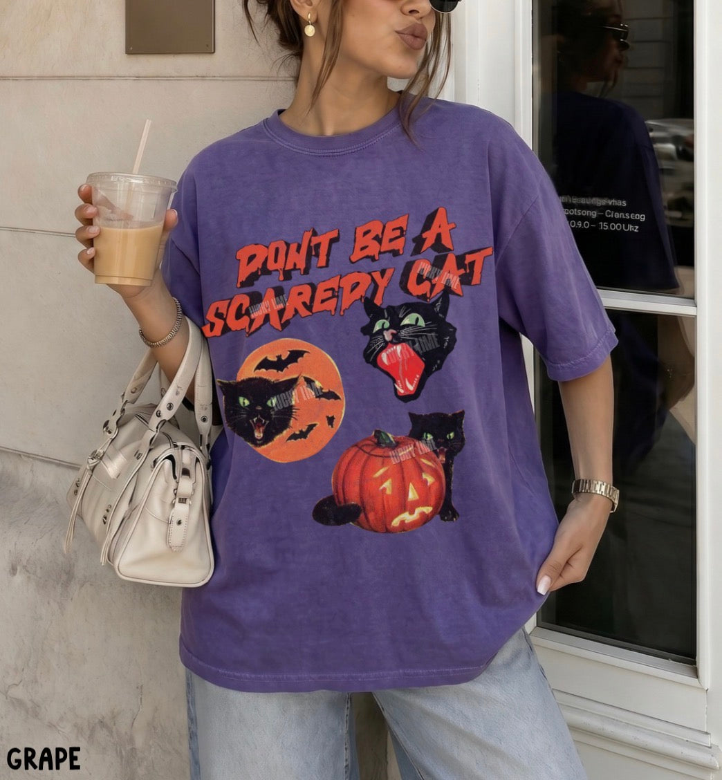 A woman wearing an oversized purple graphic T-shirt with a Halloween design that reads 'DON'T BE A SCARY CAT' above black cats, bats, a pumpkin, and an orange moon. She is holding a drink and a white handbag, standing near a storefront window.
