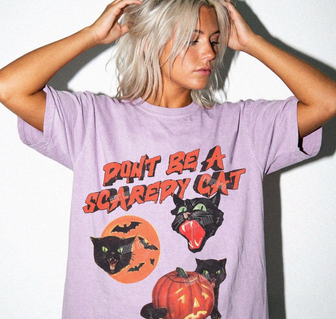 A woman with platinum blonde hair wearing a lavender graphic t-shirt that reads 'DON'T BE A SCAREY CAT' with illustrations of black cats, a jack-o'-lantern, and bats.