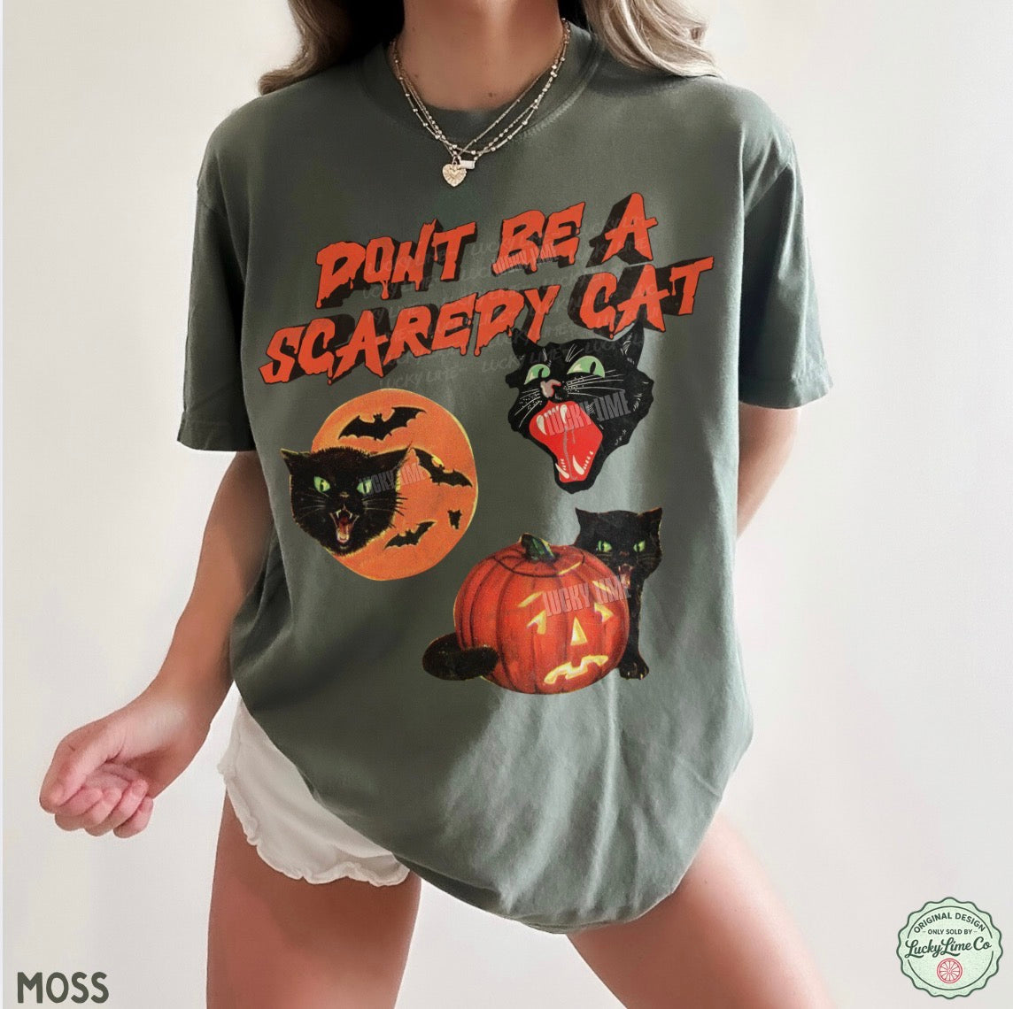 A woman wearing an oversized sage green graphic t-shirt with a Halloween theme. The shirt features the text 'DON'T BE A SCAREDY CAT' in orange, along with illustrations of a black cat in a moon, a black cat with its mouth open, and a jack-o'-lantern pumpkin.