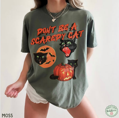 A woman wearing an oversized sage green graphic t-shirt with a Halloween theme. The shirt features the text 'DON'T BE A SCAREDY CAT' in orange, along with illustrations of a black cat in a moon, a black cat with its mouth open, and a jack-o'-lantern pumpkin.