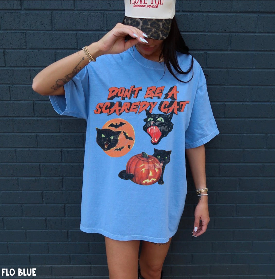 A woman wearing an oversized light blue t-shirt with a Halloween graphic that reads 'DON'T BE A SCAREDY CAT' above black cats, bats, a pumpkin, and a moon. She is posing in front of a dark gray brick wall.