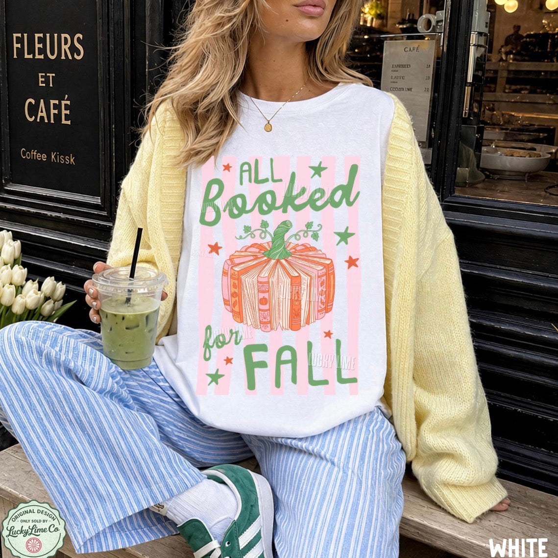 A woman sitting outdoors in front of a cafe doorway wears a white graphic T-shirt with pastel pink stripes, green text reading 'ALL Booked for FALL,' and an orange pumpkin illustration. She has on a pale yellow knit cardigan, blue-and-white striped pants, and green sneakers, and holds an iced coffee.