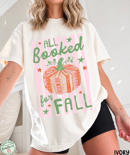 A woman wearing an oversized ivory graphic T-shirt with the phrase 'ALL Booked for FALL' in green lettering around a pumpkin illustration on a pink striped background. The photo is cropped from the neck to the upper thighs, with black shorts visible.