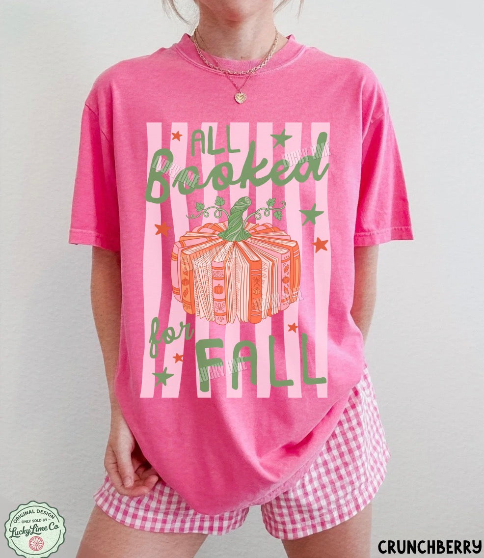 A person wearing a bright pink graphic T-shirt with the words 'ALL Booked for FALL' and a pumpkin illustration, paired with pink-and-white gingham shorts against a plain light background.
