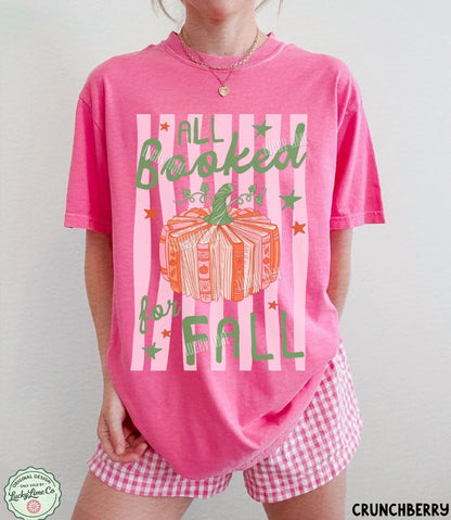 A person wearing a bright pink graphic T-shirt with the words 'ALL Booked for FALL' and a pumpkin illustration, paired with pink-and-white gingham shorts against a plain light background.