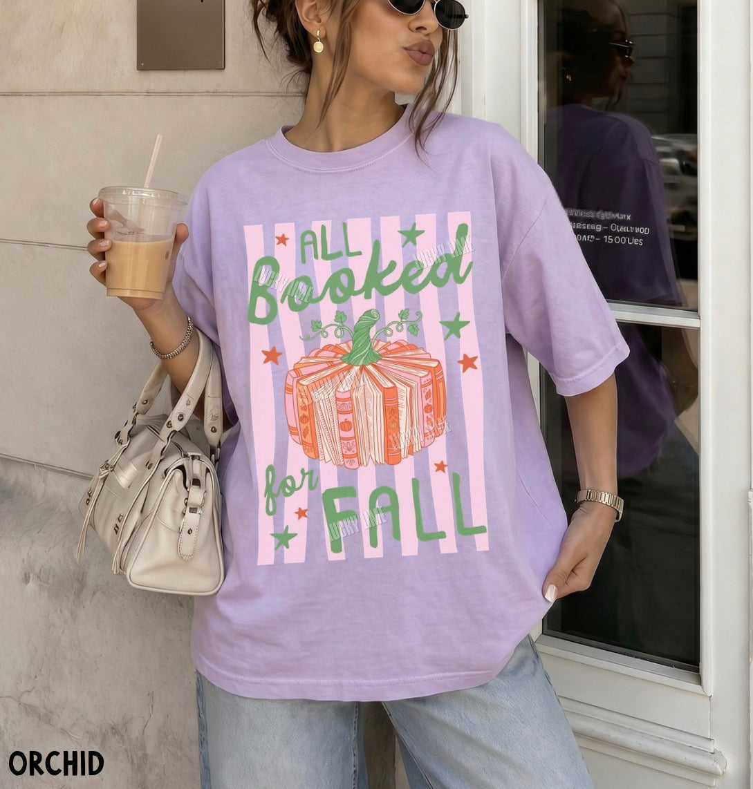 A woman wearing an oversized lavender graphic T-shirt with a pink striped design, green text reading 'ALL Booked for FALL,' and an orange wrapped gift illustration, standing outdoors near a window and holding an iced coffee.