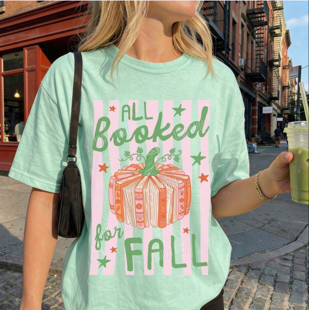 A woman wearing a mint green graphic t-shirt with a fall-themed book club design that reads 'ALL Booked for FALL' with an illustration of a wrapped gift box. She is holding an iced coffee on a cobblestone street.