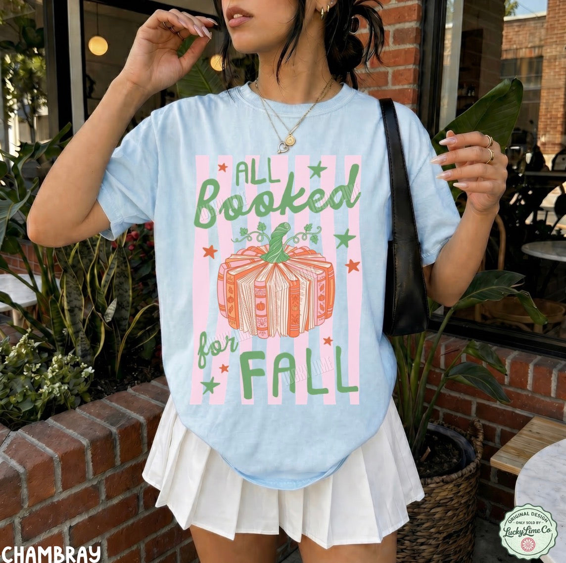 A woman wearing a light blue graphic t-shirt with a 'All Booked for Fall' autumn pumpkin design, styled with a white pleated skirt in an outdoor patio setting.