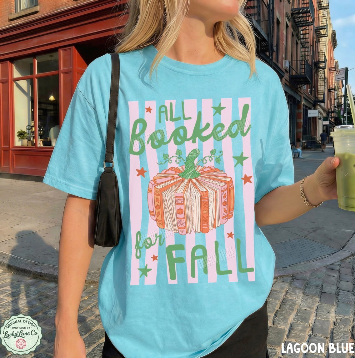 A woman wearing a light blue graphic t-shirt with a fall-themed book-themed design that reads 'ALL Booked for FALL' with an illustration of a wrapped book. She is holding a green drink and wearing a black shoulder bag, set against a blurred street background.