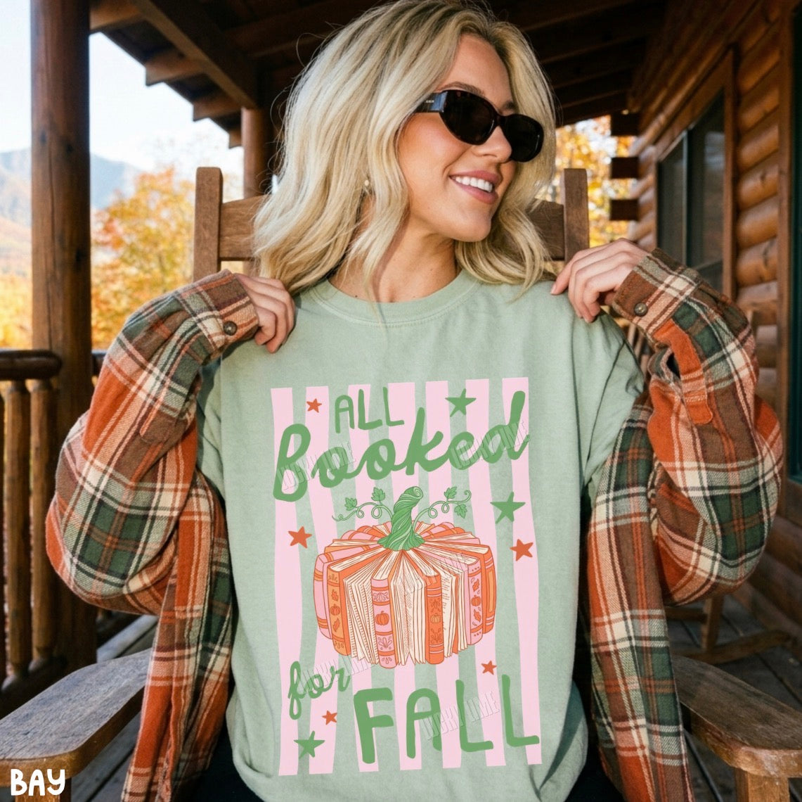 A woman wearing a sage green graphic t-shirt with a fall-themed design, styled with a rust-orange plaid flannel shirt on a rustic wooden porch.