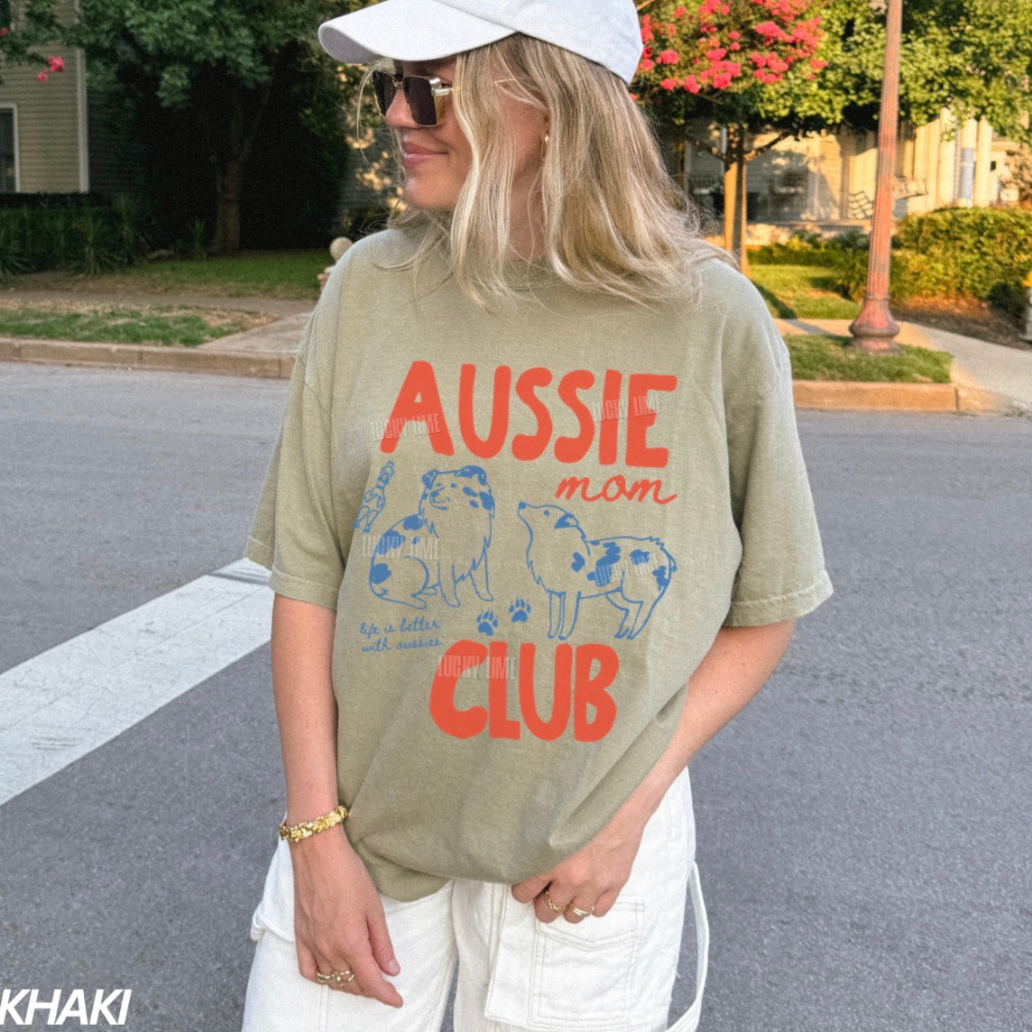 A woman wearing a khaki graphic t-shirt with 'AUSSIE mom CLUB' printed in orange and blue, paired with white pants and a white cap, standing on a suburban street.