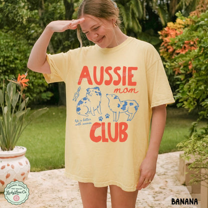 A woman wearing a pale yellow graphic T-shirt outdoors in a garden setting. The shirt features the words 'AUSSIE mom CLUB' in red and blue, with illustrations of two dogs.