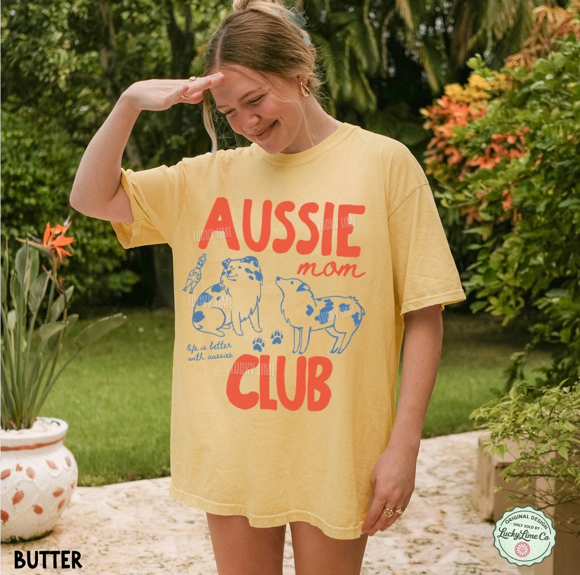 A woman wearing a yellow graphic t-shirt with 'AUSSIE mom CLUB' printed in red and blue, standing outdoors in a garden setting.